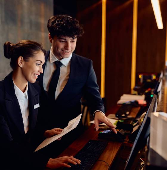 Happy hotel managers through reservation list on desktop PC at reception desk.